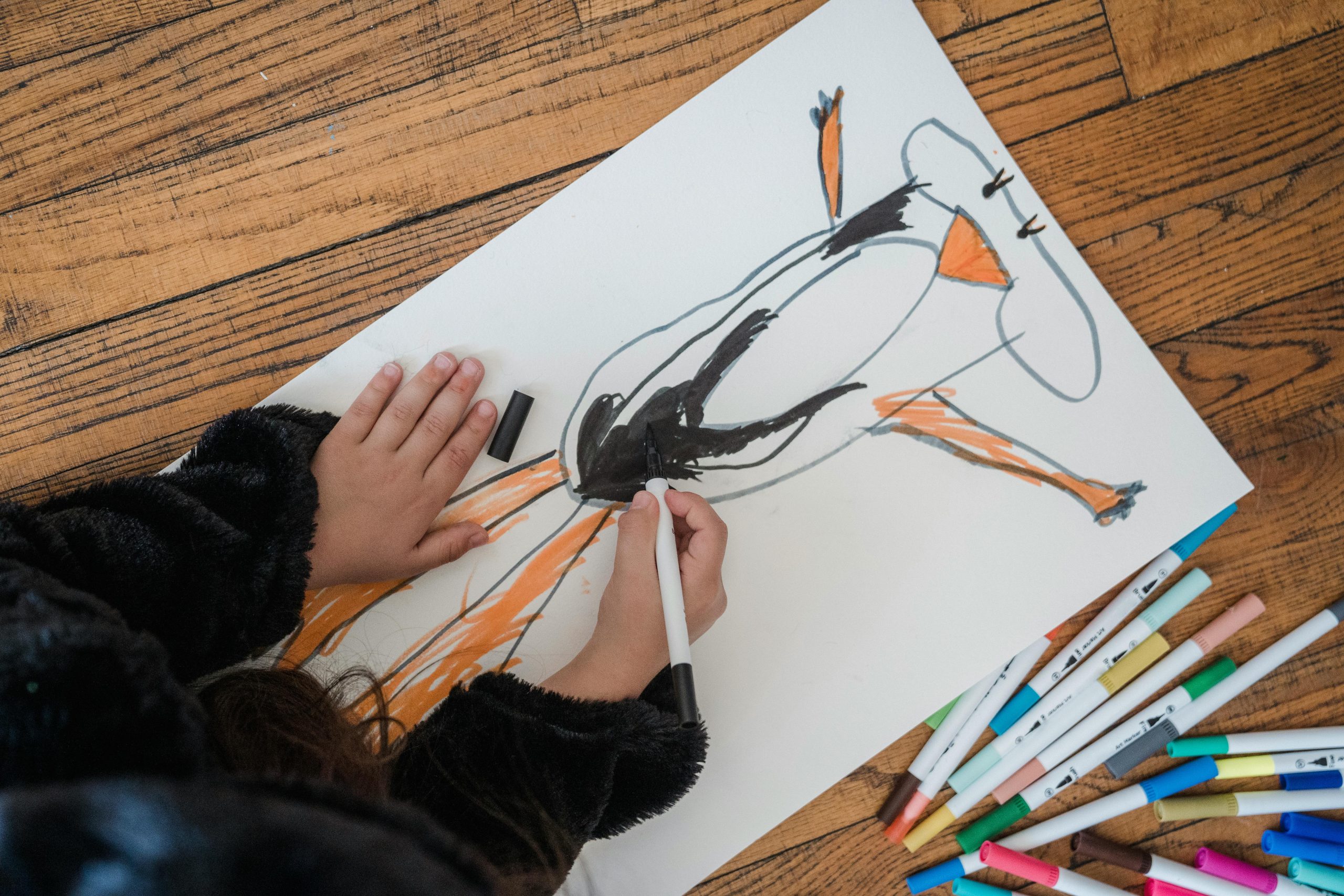 A child drawing a colorful picture on paper using various markers placed on a wooden surface.